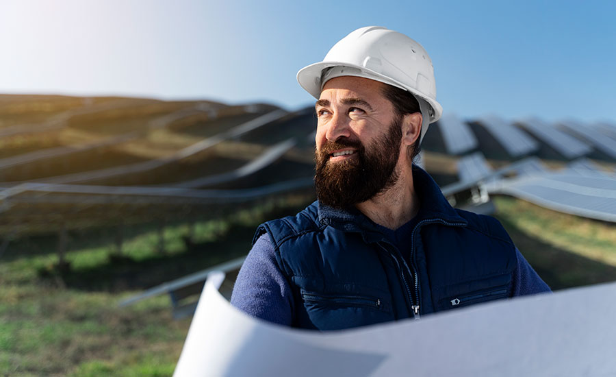 A bearded man in a white hard hat and blue vest holds blueprints, standing before a large solar panel farm on a sunny day, looking thoughtful.