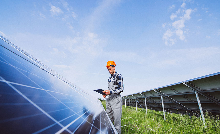 A worker in a checkered shirt and hard hat inspects solar panels with a tablet in hand.