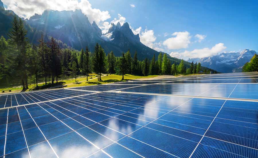 A field of solar panels under a bright blue sky near a forest with tall, jagged mountains in the background.