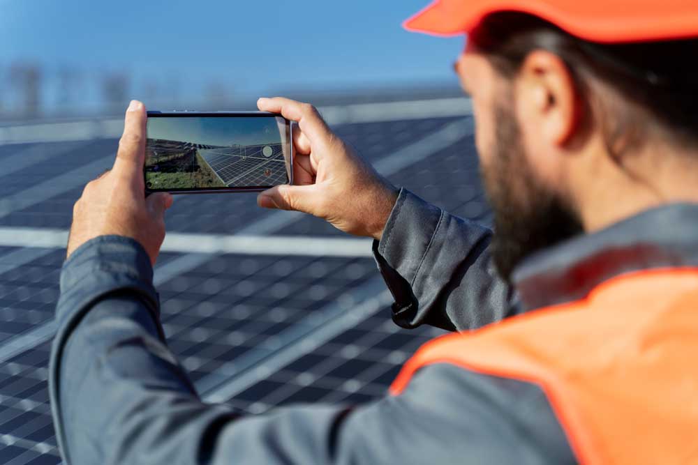 Person in an orange vest and cap takes a photo of solar panels under a clear blue sky.