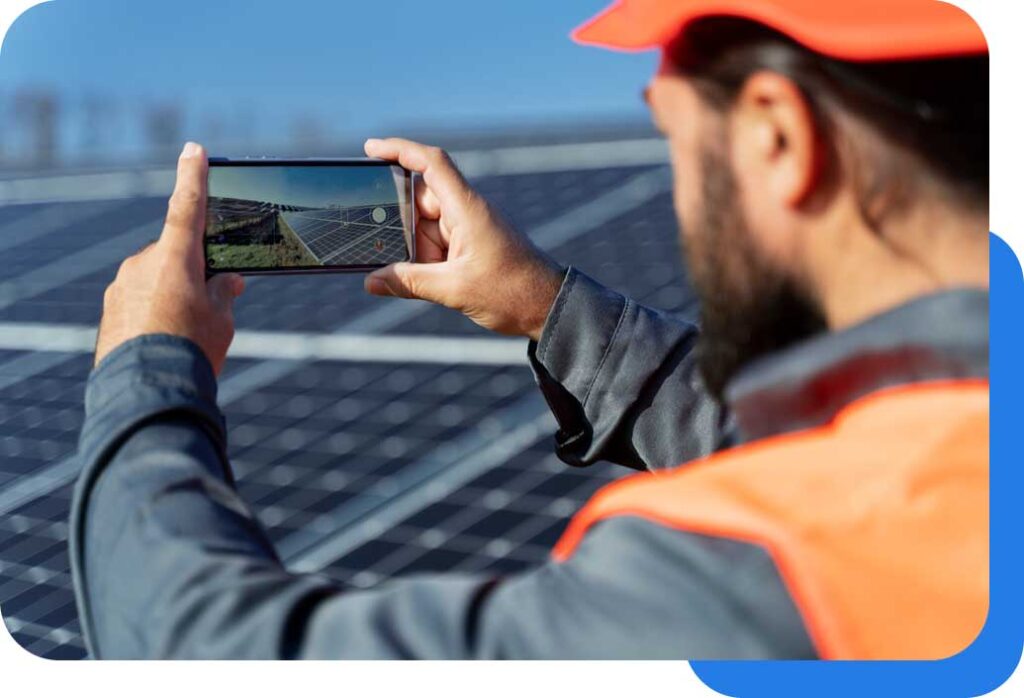 A man in an orange safety vest and helmet uses his smartphone to take a photo of a solar panel installation on a sunny day.