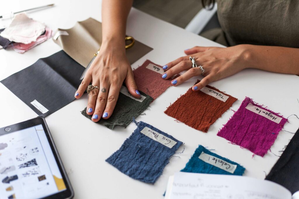 A person with rings and blue nail polish selects labeled fabric swatches on a white table with a tablet and notebook.