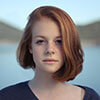 A young person with red hair and a neutral expression is close-up, with a blurred background of water and hills under a clear sky.