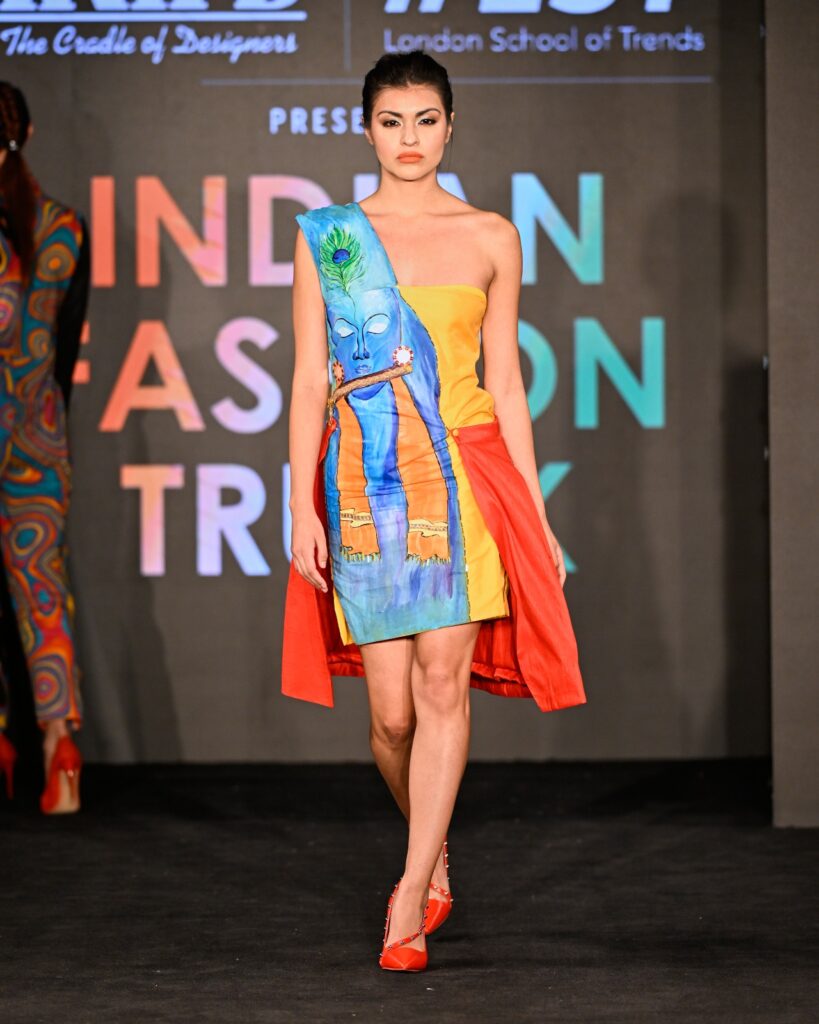 A model in a colorful asymmetrical dress with a red cape walks the runway. The backdrop says Indian Fashion Trunk and London School of Trends.