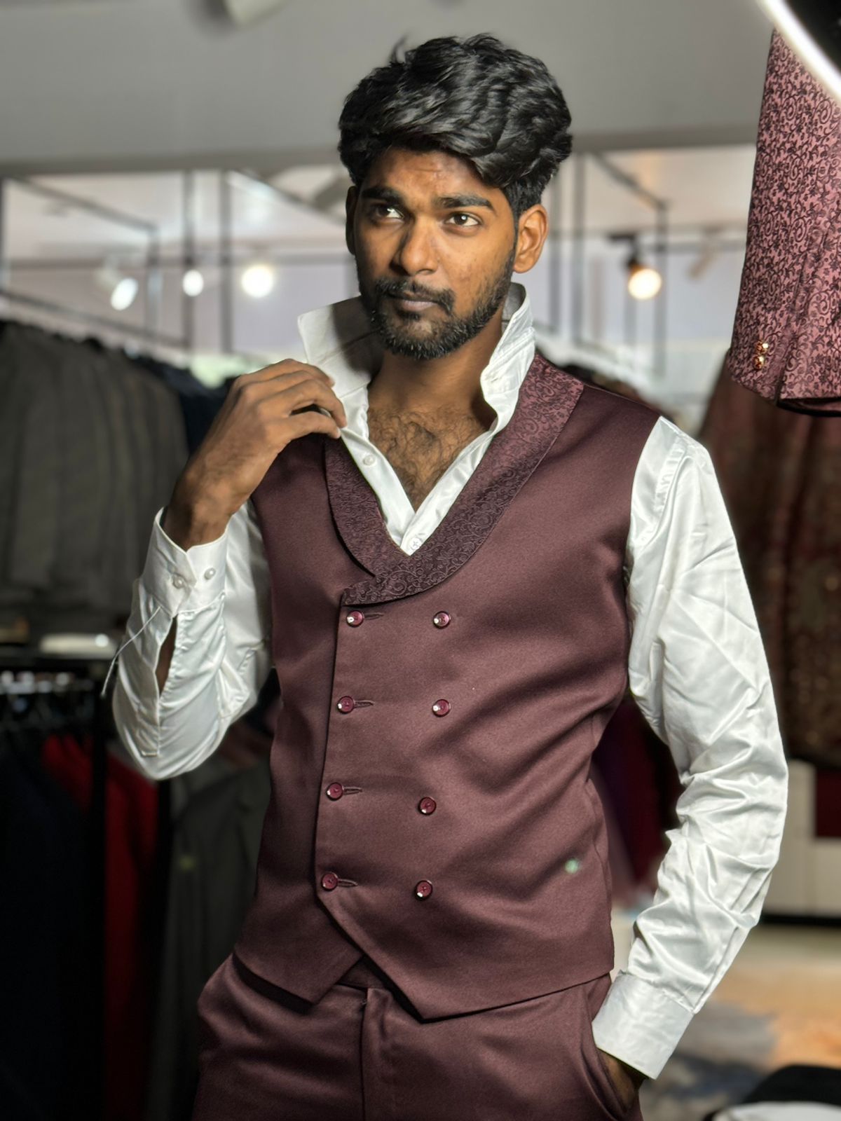 A man with dark hair and beard wears a maroon double-breasted vest over a white dress shirt, posing indoors.