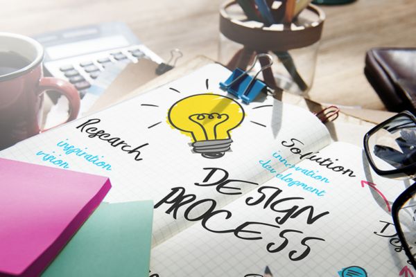 A workspace with a DESIGN PROCESS notebook, light bulb drawing, notes Research and Solution, and scattered office supplies.