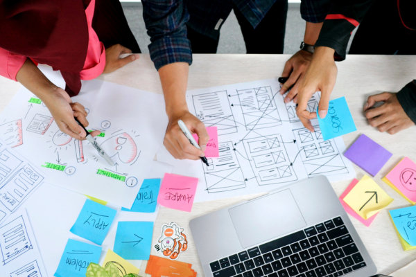 Four people collaborate at a workspace with wireframes, sticky notes, and sketches, discussing and designing.